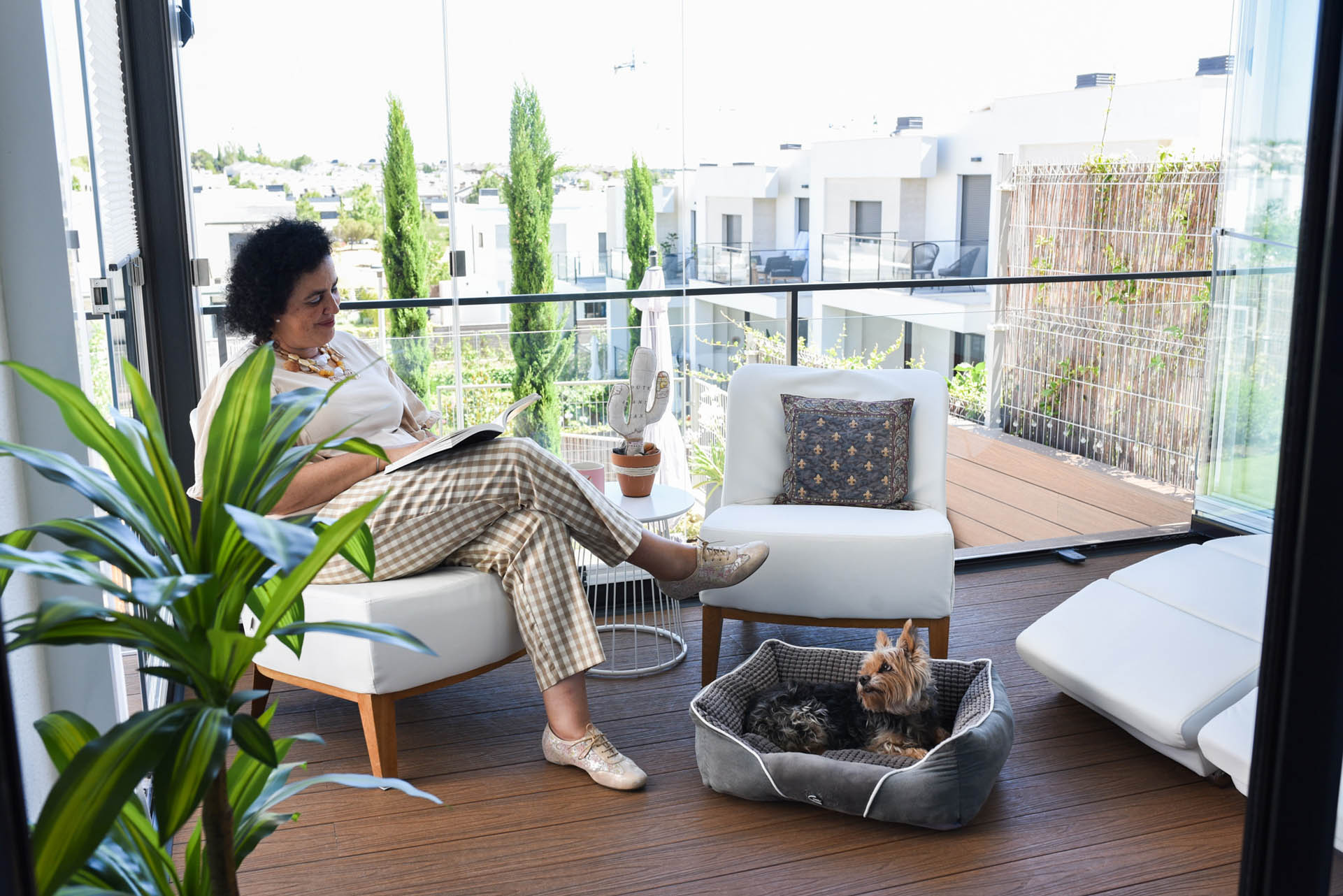 Eine Frau sitzt auf ihrem Balkon und liest. Vor ihr sitzt ein Hund. Die Balkonverglasung ist zum Teil geöffnet. Man sieht die Balkone der Nachbarschaft.