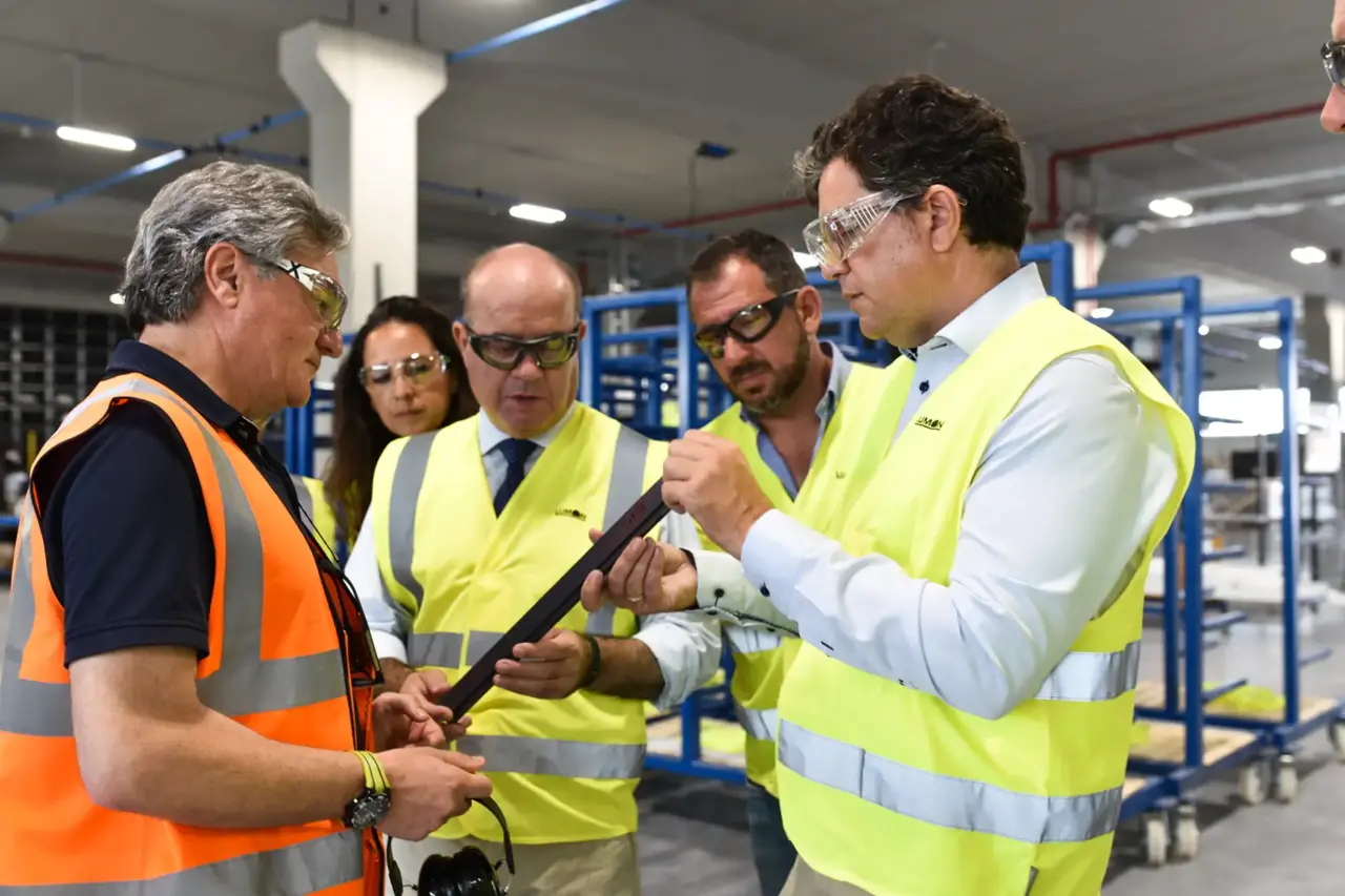 Eine Frau und drei Männer mit Warnwesten und Schutzbrillen begutachten ein Glasprofil für eine Balkonverglasung. Im Hintergrund stehen die Produktionsanlagen.