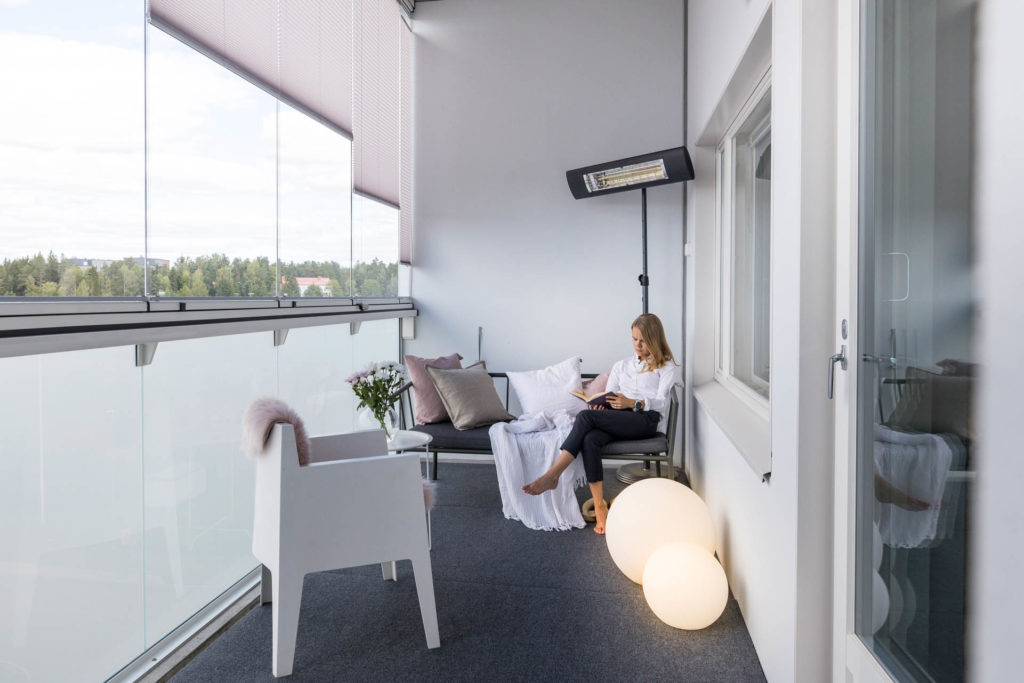 Eine Frau sitzt auf einer Bank auf ihrem Balkon und liest. Durch die Verglasung bestehend aus einem Ganzglasgeländer und einer rahmenlosen Balkonverglasung ist sie vor der Kälte geschützt. Hinter ihr steht ein Heizstrahler.