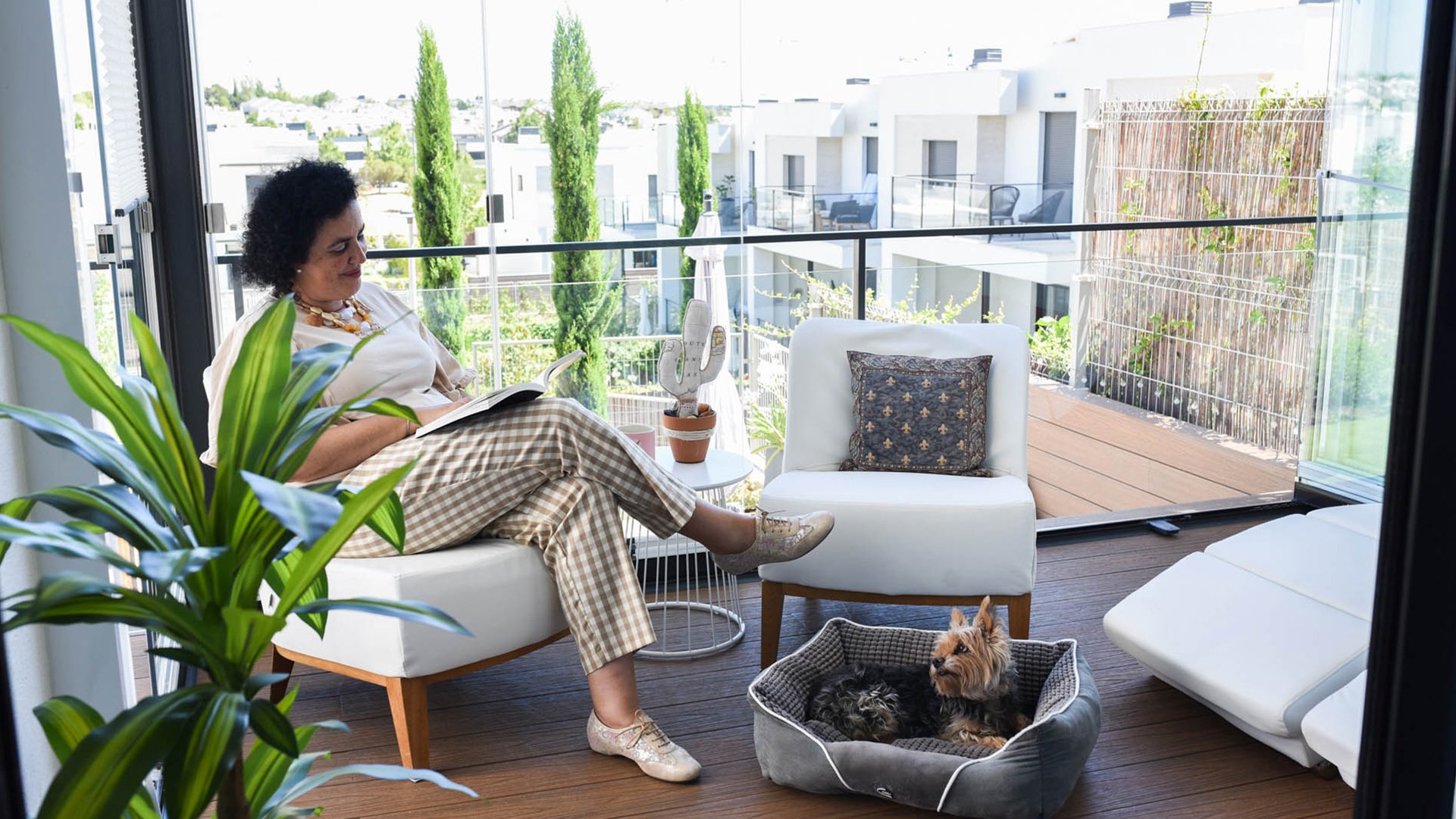 Eine Frau sitzt auf ihrem Balkon und liest. Vor ihr sitzt ein Hund. Die Balkonverglasung ist zum Teil geöffnet. Man sieht die Balkone der Nachbarschaft.