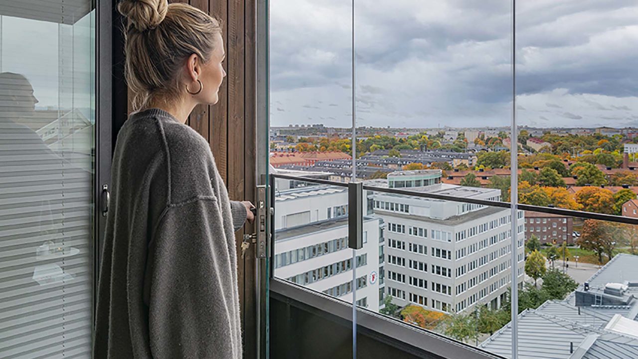 Eine Frau öffnet ihre Balkonverglasung. Im Hintergrund ist eine Stadt zu sehen.