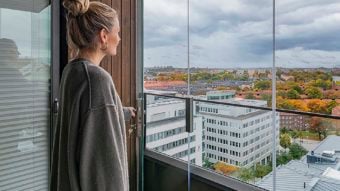 Eine Frau öffnet ihre Balkonverglasung. Im Hintergrund ist eine Stadt zu sehen.