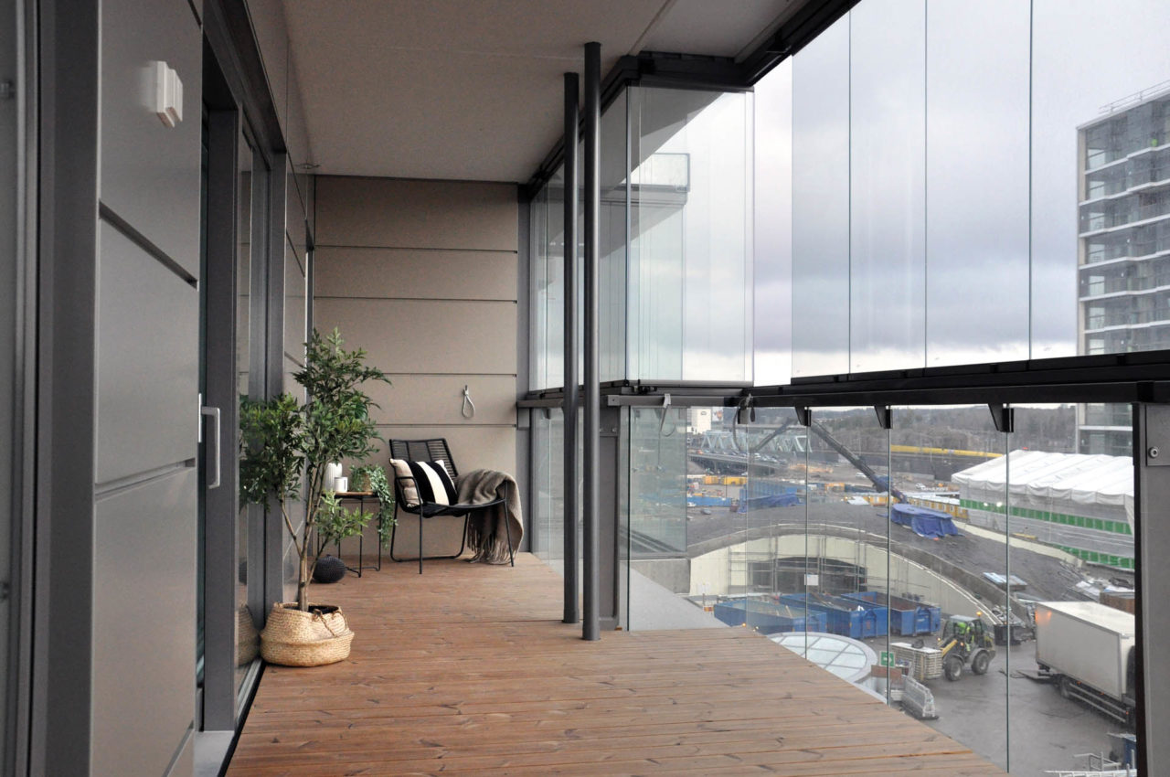 Blick auf einen Balkon von innen mit Holzboden, ein paar Pflanzen und einer Dreh/Schiebeverglasung auf einem Ganzglasgeländer aufgesetzt.