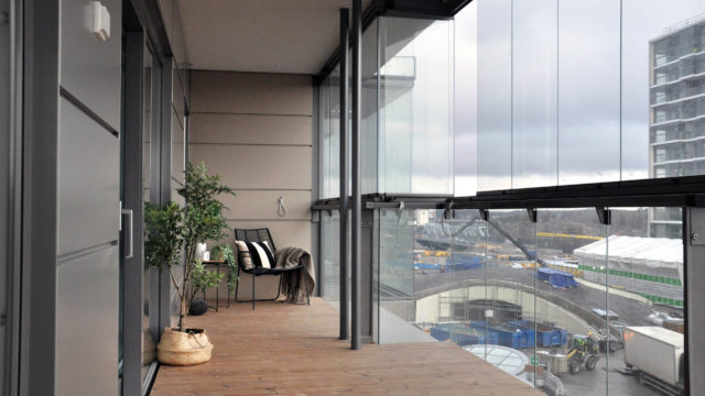 Blick auf einen Balkon von innen mit Holzboden, ein paar Pflanzen und einer Dreh/Schiebeverglasung auf einem Ganzglasgeländer aufgesetzt.