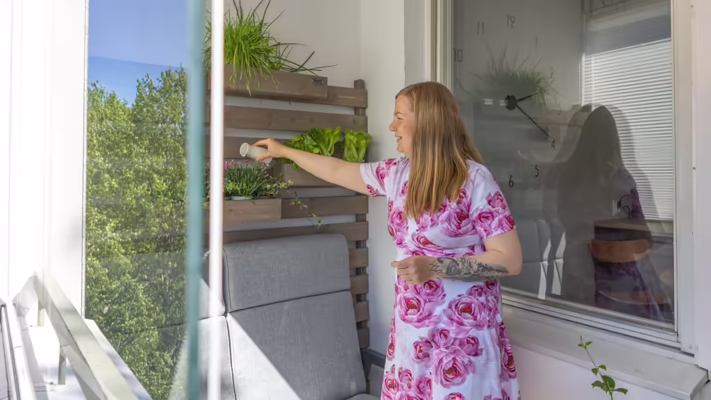 Blick von draußen auf einen verglasten Balkon auf dem eine Frau in pinkem Kleid ihre Pflanzen gießt.