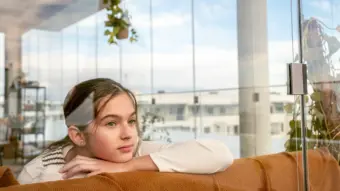 Ein Mädchen träumt vor sich hin. Sie stützt ihr Kinn auf einer Sofa-Lehne ab. Im Hintergrund sieht man eine geschlossene Balkonverglasung