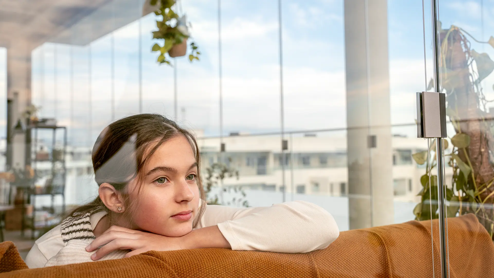 Ein Mädchen träumt vor sich hin. Sie stützt ihr Kinn auf einer Sofa-Lehne ab. Im Hintergrund sieht man eine geschlossene Balkonverglasung